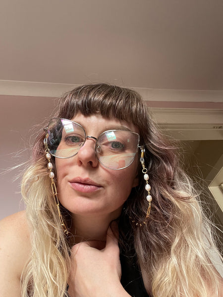 Three pearls hang from either side of the gold glasses chain. A woman is wearing oversized cat eye glasses along with the beautiful pearl and gold glasses chain. 