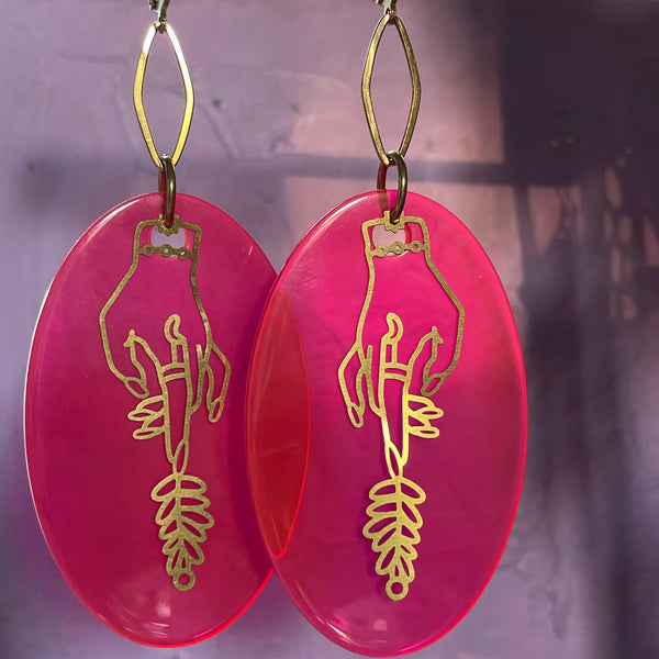 Long oval acrylic in hot pink with a delicate brass hand layered on top. The brass hand has a moon ring and is holding some herbs/plants. The hot pink and gold earrings hang from a hollow diamond shaped connecter and have leverback ear wires. The lever back earwires are stainless steel. The hot pink oval is translucent yet very vibrant. 
